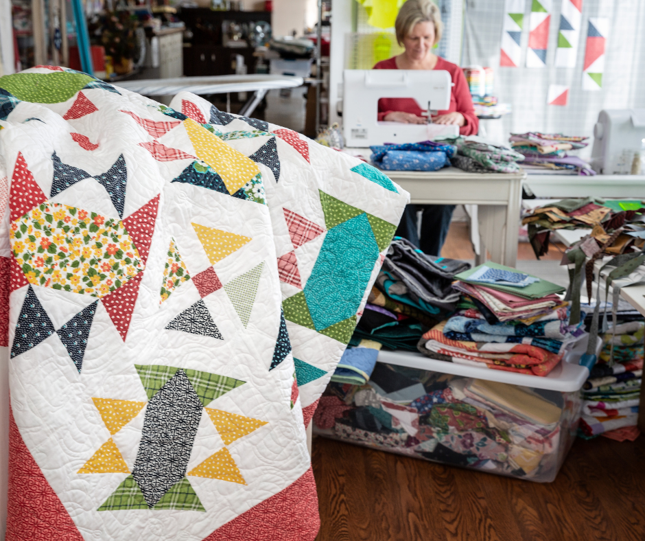 a colorful quilt in a sewing studio with a woman sewing on a machine in the background.