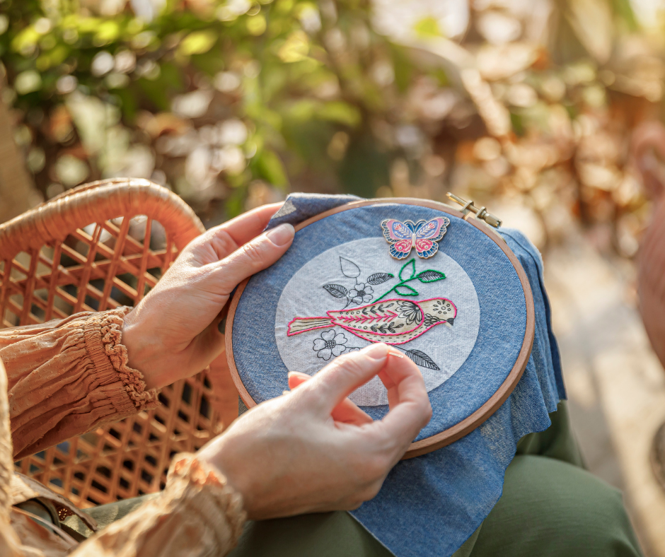 Hands hold an embroidery hoop with a colorful bird and butterfly design, set against a blurred, sunlit garden background.