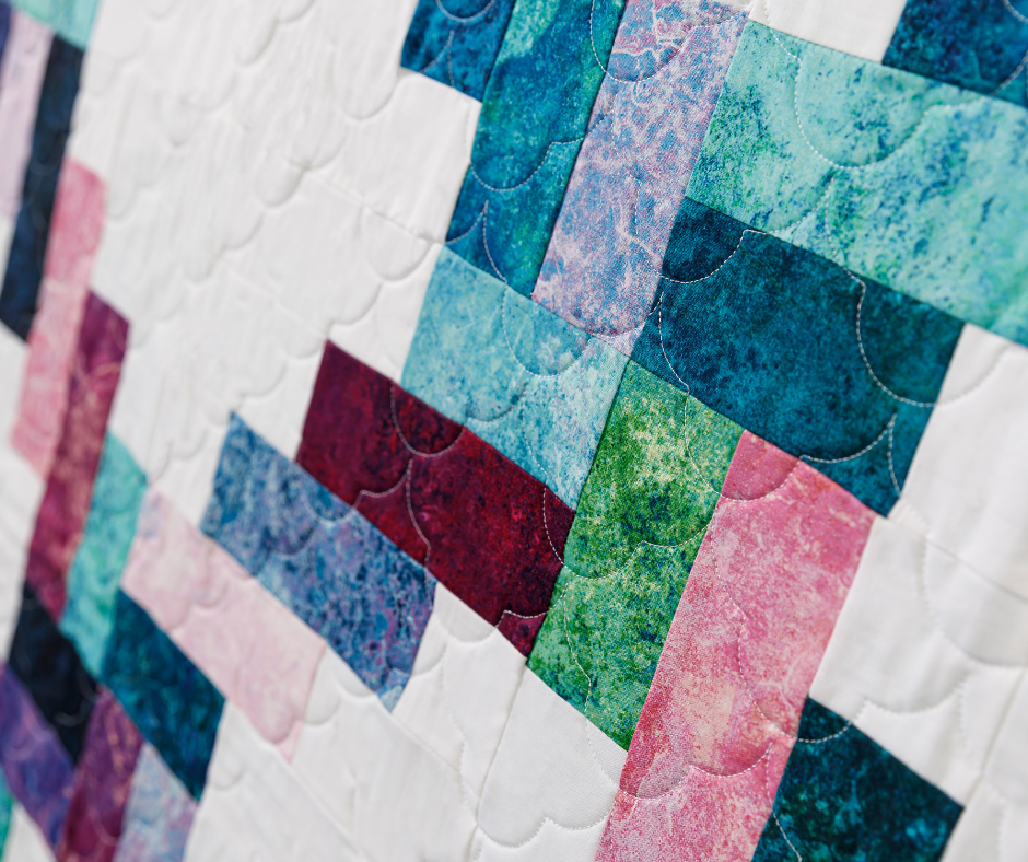 Close-up of a colorful quilt with interlocking geometric blocks. The vibrant marbled fabric patterns include shades of teal, blue, red, and pink, conveying warmth.