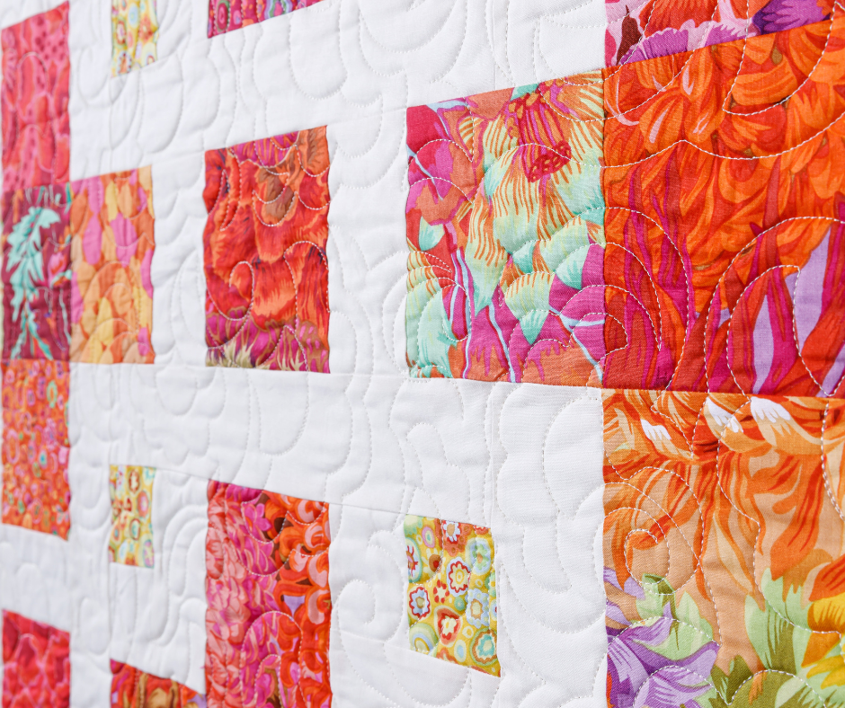 Colorful quilt pattern with vibrant square patches of floral and abstract designs in reds, oranges, and pinks on a white background.