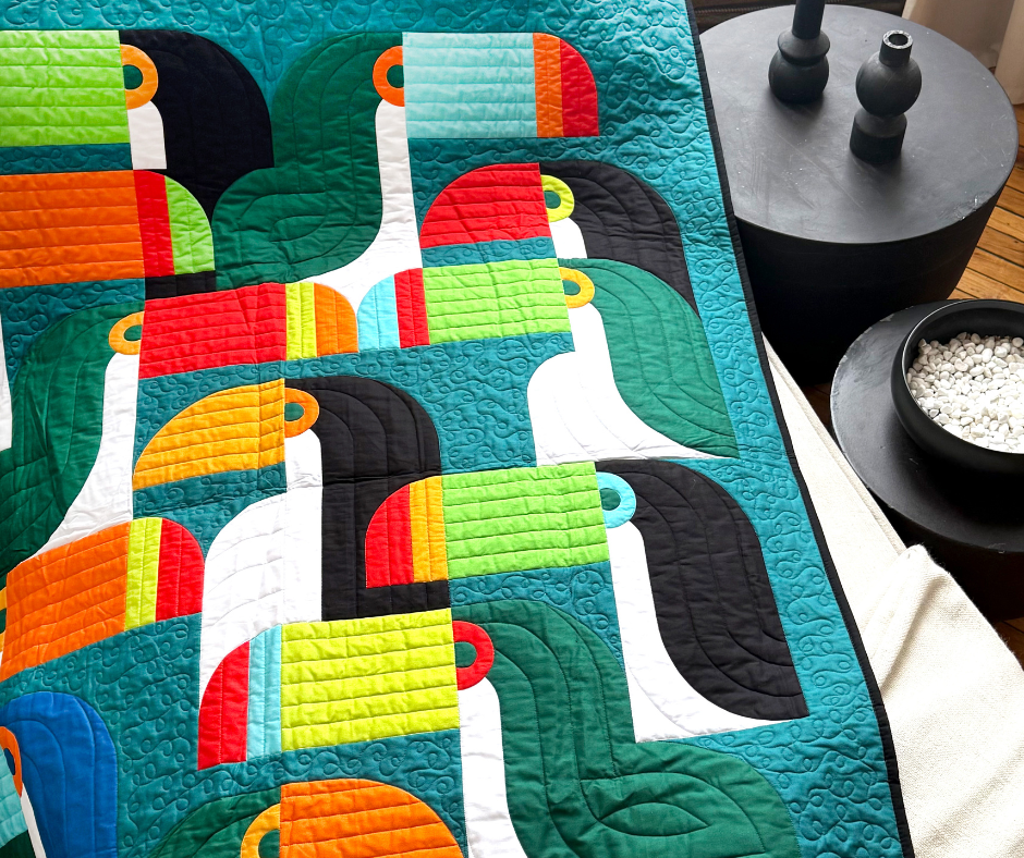 Colorful quilt with toucan patterns in black, green, orange, and red against a teal background. The quilt rests on furniture with a modern, cozy vibe.